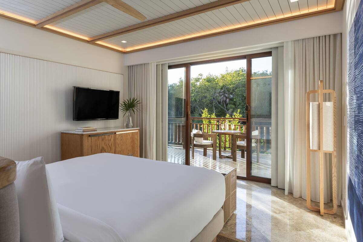 A modern hotel room with a large bed, a flat-screen TV, a balcony with outdoor seating, and a view of greenery.