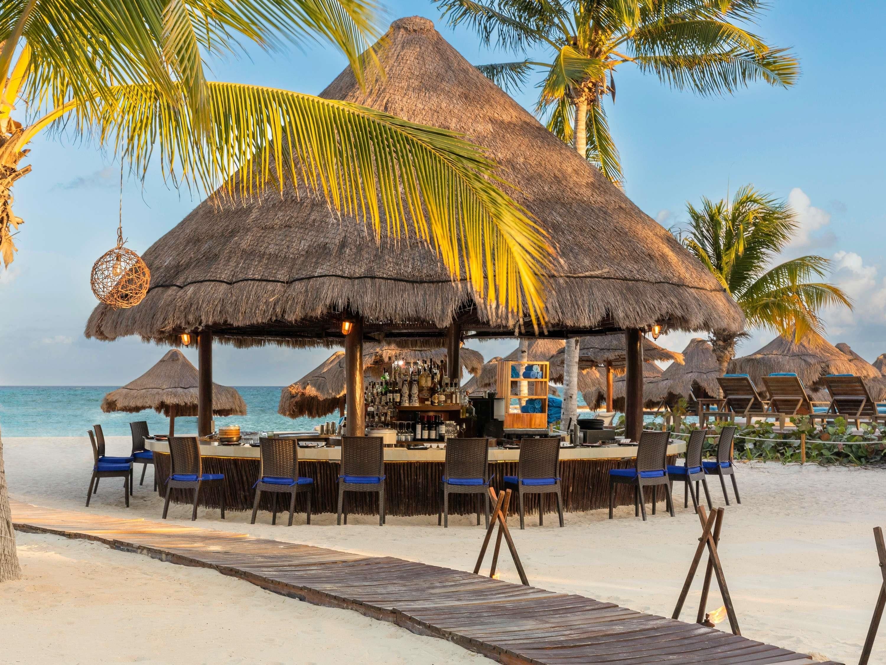 Eine Strandbar mit reetgedeckten Sonnenschirmen, Liegestühlen und einem Holzdeck, das zum Sand führt.