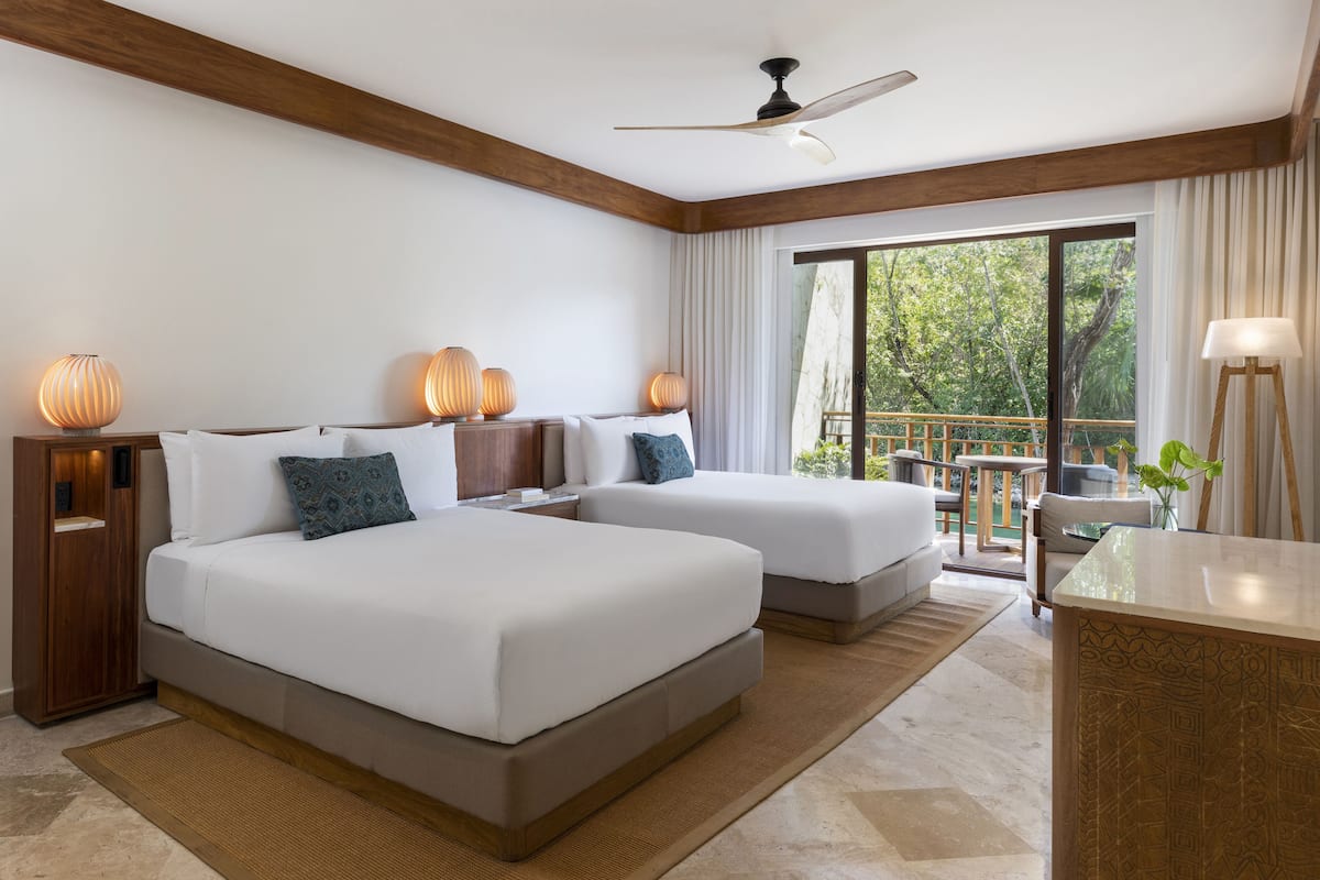 A hotel room with two beds, a desk, a chair, a lamp, and a view of the outdoors.