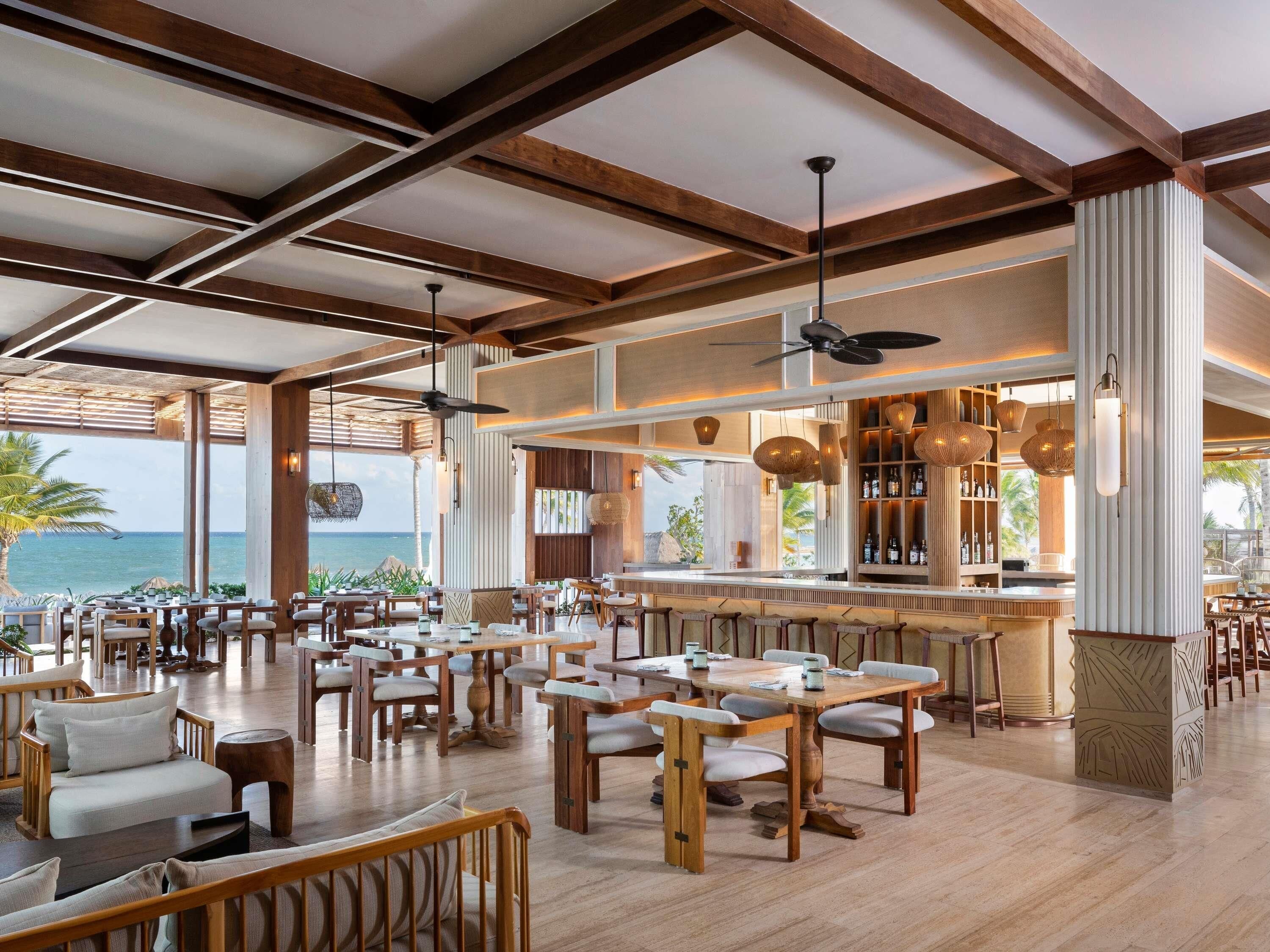 A spacious restaurant with wooden furniture, a bar area, and large windows offering a view of the ocean.