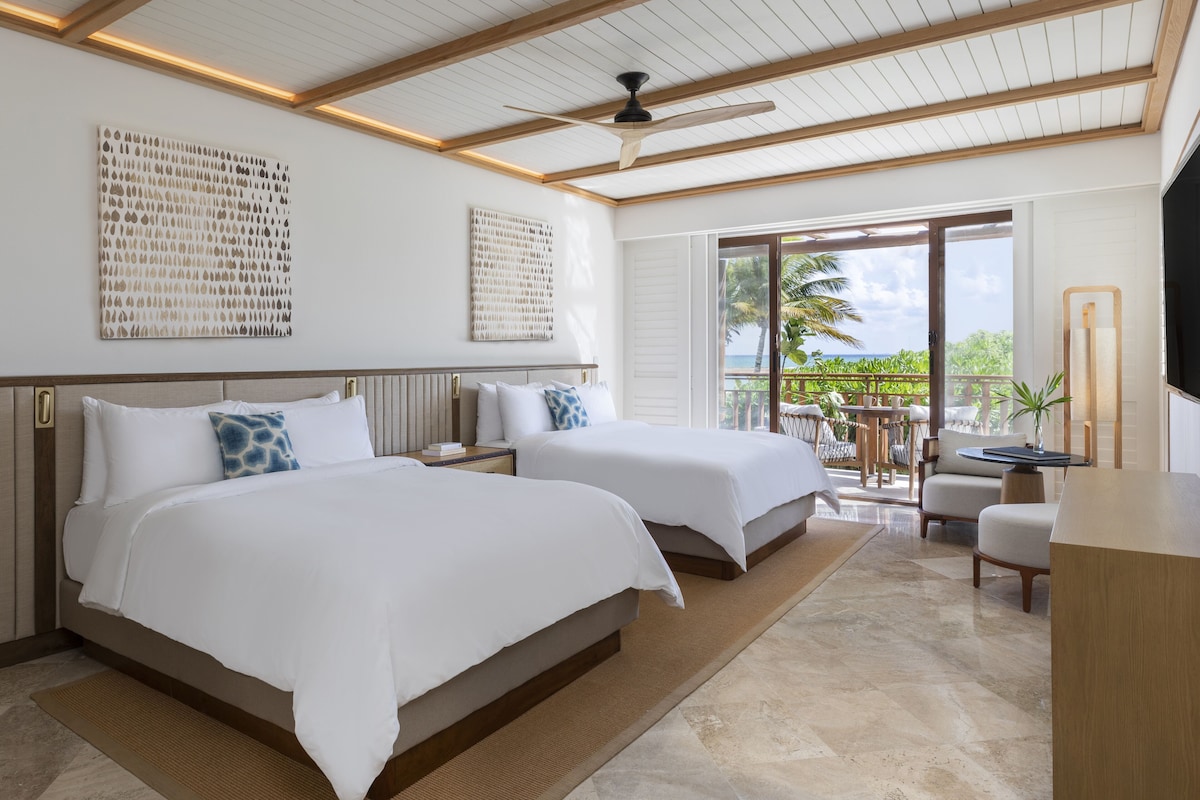 Une chambre d’hôtel avec deux lits, un ventilateur de plafond, des œuvres d’art accrochées aux murs, un balcon avec vue sur l’océan et un téléviseur situé sur le côté droit.
