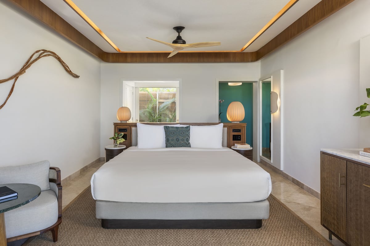 A modern hotel room with a large bed, wooden ceiling, and a view of greenery outside.