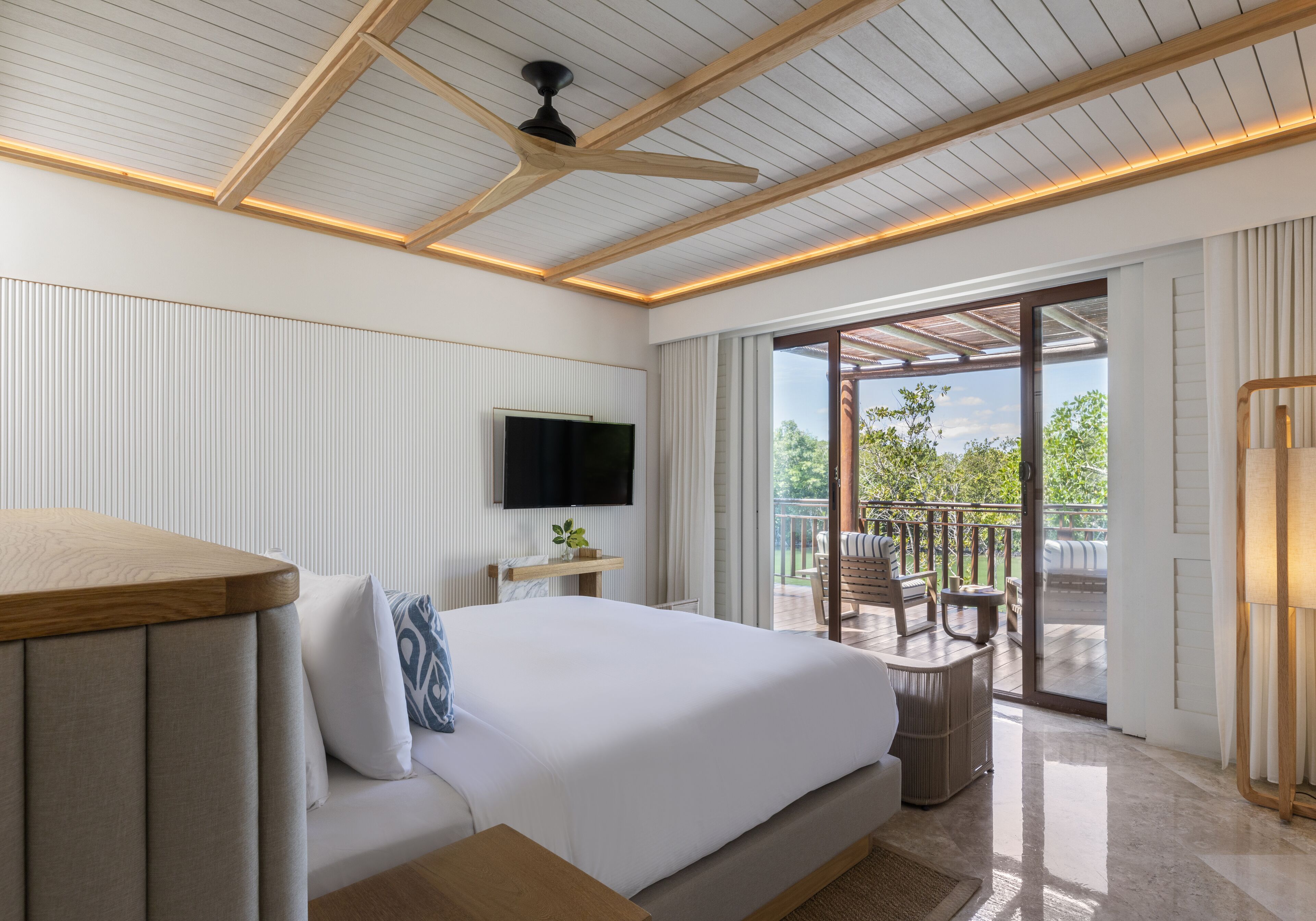 A modern bedroom with a large bed, a TV, a wooden ceiling, and a balcony with outdoor furniture.