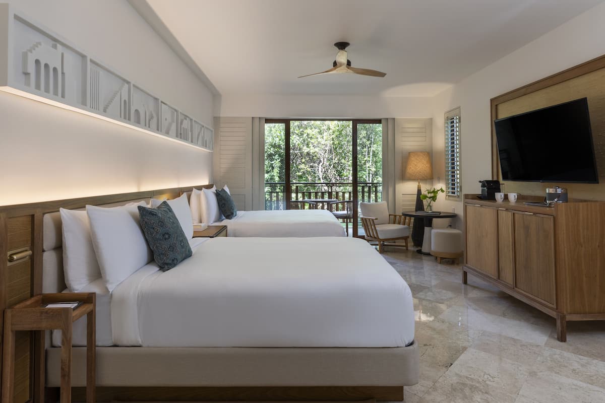 A hotel room with two beds, a TV, a desk, and a balcony with a view of trees.