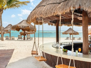 Un bar situé en bord de mer, doté de parasols en chaume, de chaises longues et offrant une vue sur l’océan.