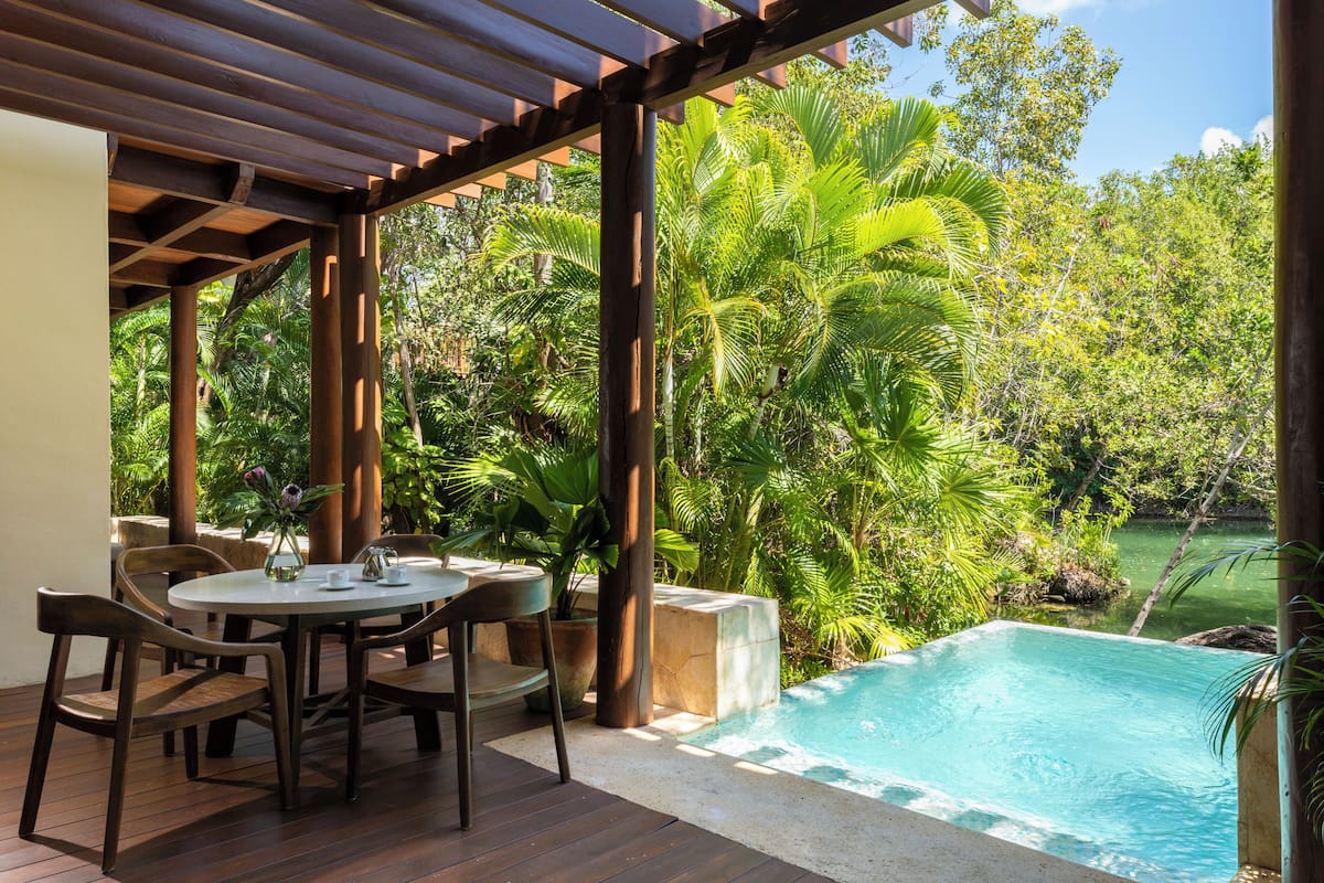 A covered outdoor area with a pool, wooden furniture, and lush greenery.