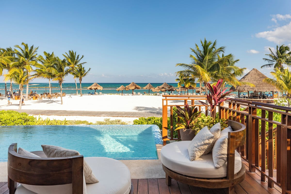 A poolside area with a view of the beach, lounge chairs, and a thatched-roof structure.