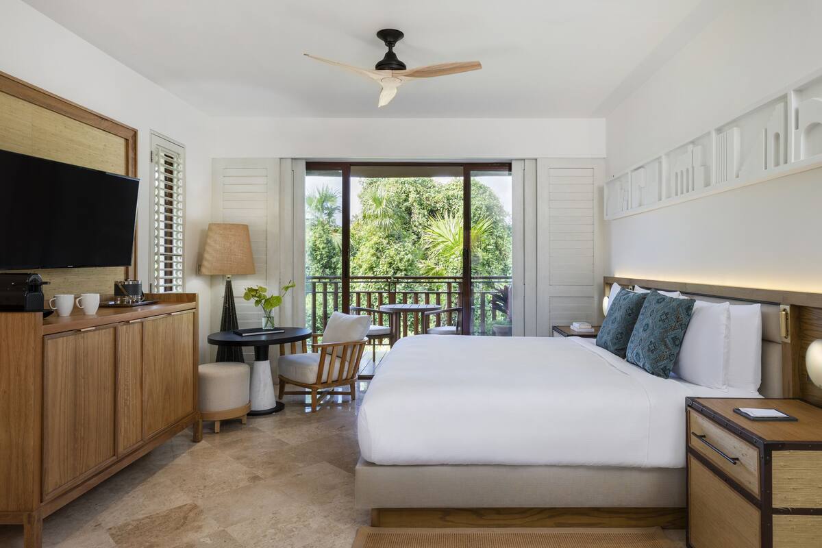 A hotel room with a bed, a desk, a television, a balcony with a view of greenery.