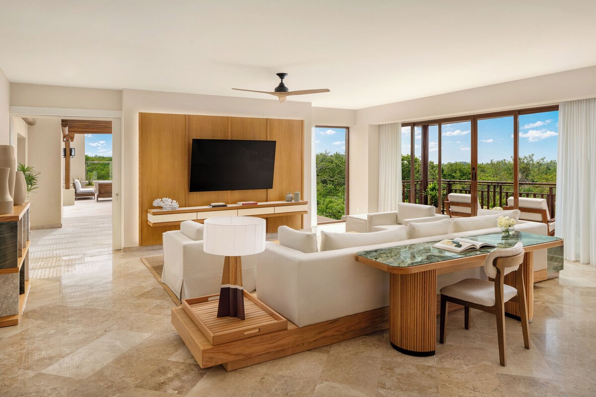 A modern living room with a large flat-screen TV, a wooden entertainment unit, a glass coffee table, and a seating area with white sofas and a dining table.