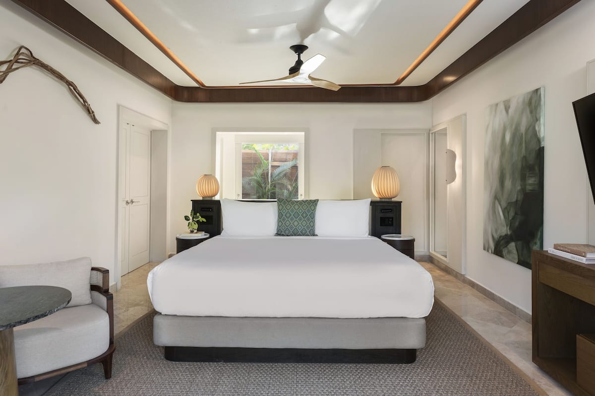 A modern hotel room with a large bed, two bedside tables with lamps, a ceiling fan, a window with a view of greenery, and a flat-screen TV on a wooden console.