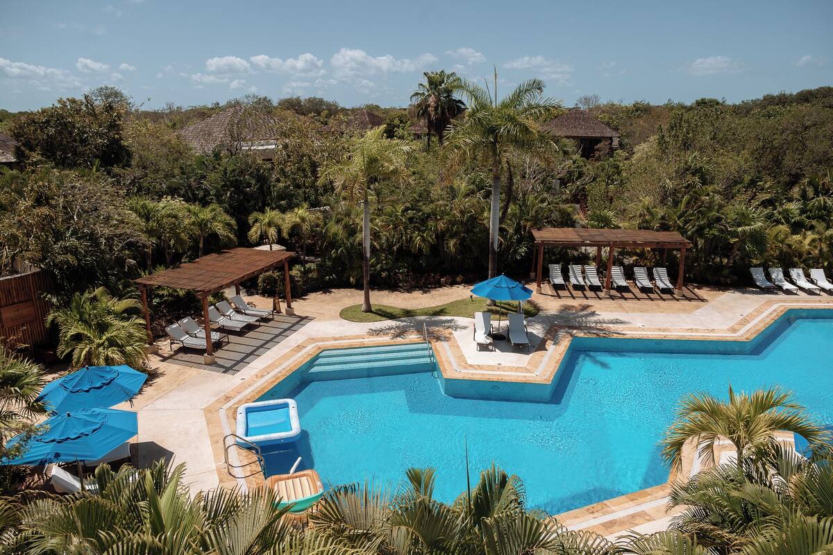 A swimming pool with lounge chairs and umbrellas, surrounded by palm trees and a hilly landscape.