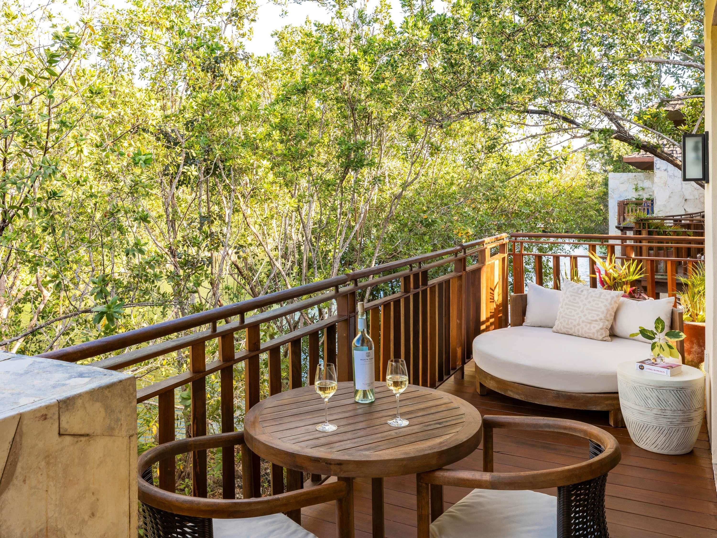 A wooden deck with a round table, a cushioned seating area, and a view of dense foliage.