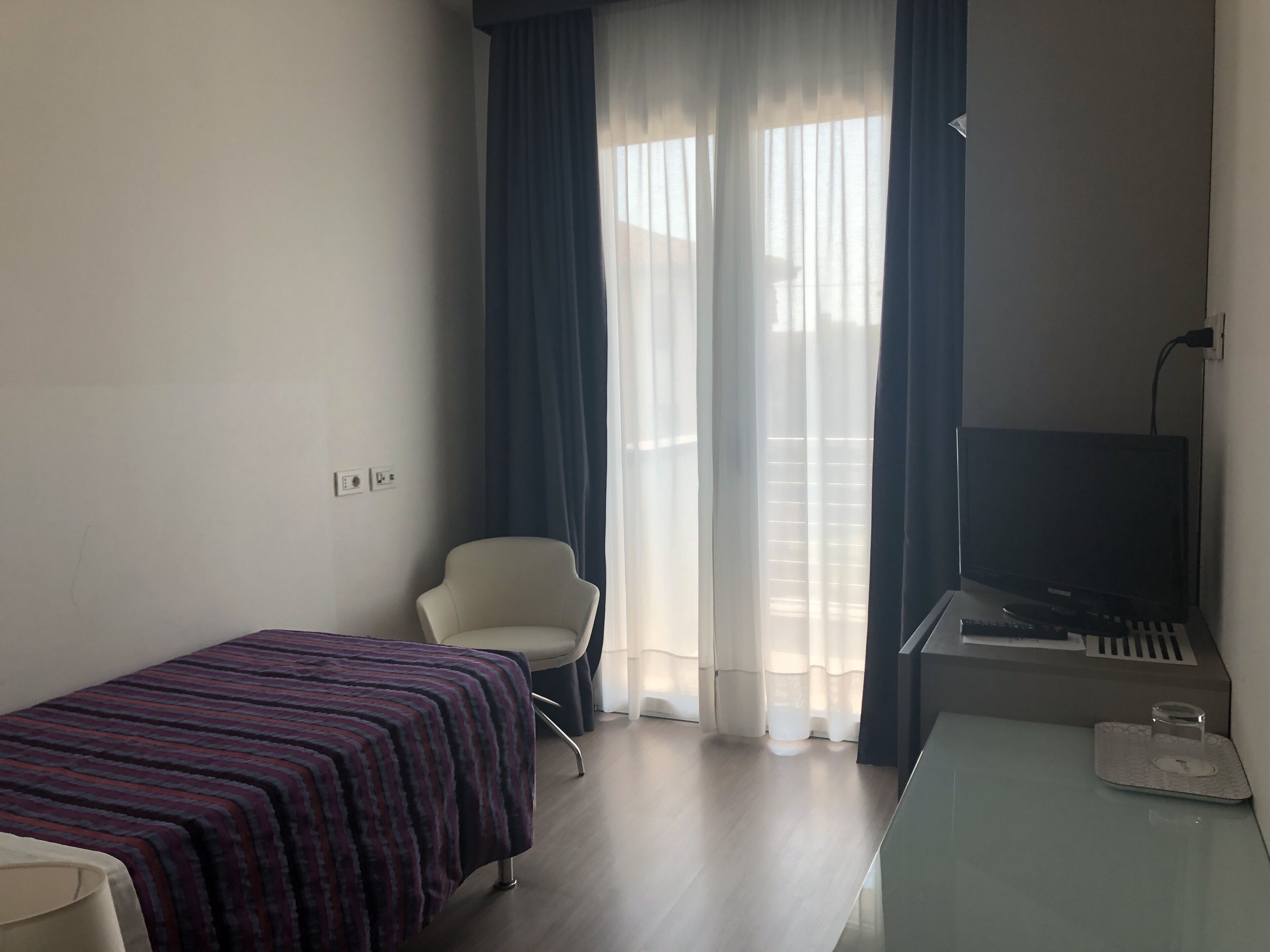 A hotel room with a bed, a chair, a desk with a keyboard, a TV, and a window with curtains.