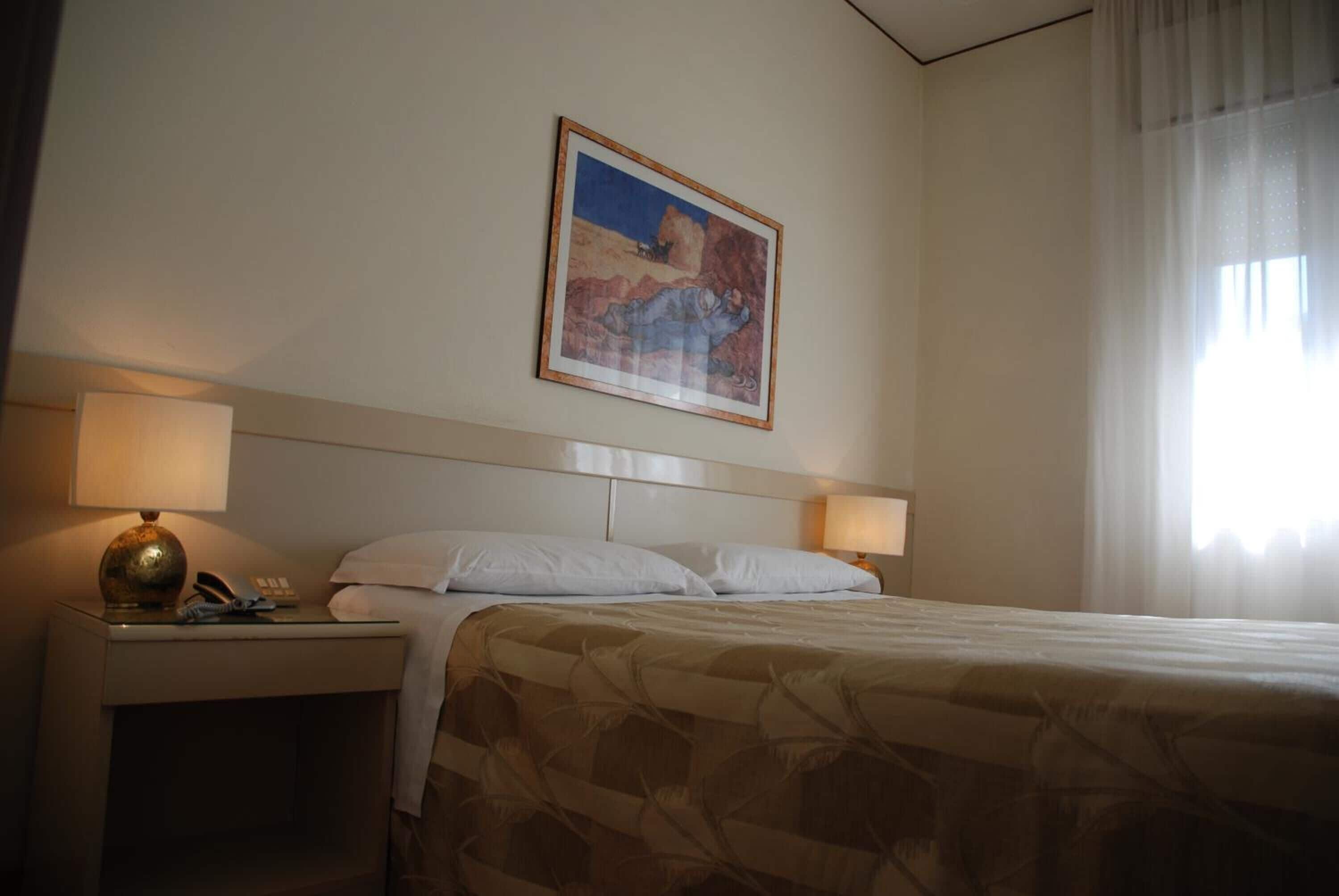 Ein Hotelzimmer mit einem Bett, zwei Nachttischlampen, einem Nachttisch mit Telefon und einem gerahmten Bild an der Wand.