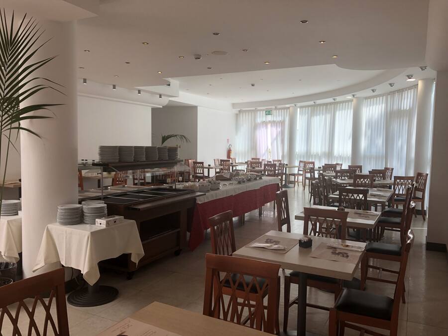 Un restaurante con mesas, sillas, una zona de bufé y amplias ventanas con cortinas.