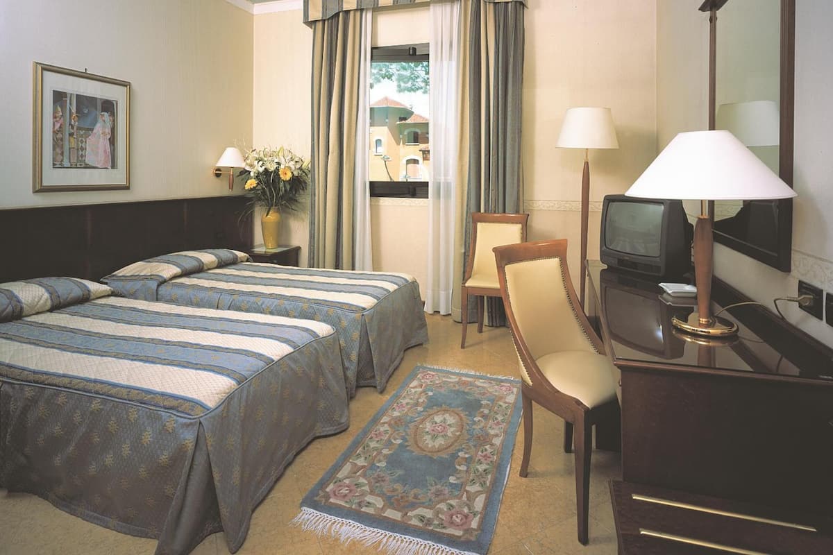 Une chambre d’hôtel avec deux lits, un bureau avec une télévision, une chaise, une lampe de bureau et une fenêtre avec des rideaux.
