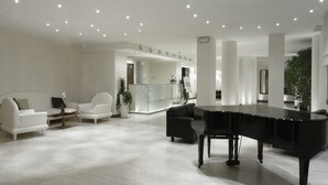 A spacious living room with a grand piano, white sofas, and a reception area in the background. - Hotel Lugano Torretta (Mestre)