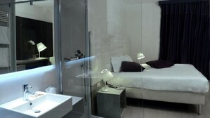 A modern hotel room with a glass shower enclosure, a bedside table with a lamp, a bed with pillows, and a large mirror above the sink. - Hotel Lugano Torretta (Mestre)