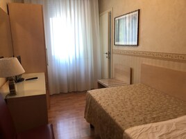 Una camera d'albergo con un letto, una scrivania con lampada, uno specchio e una finestra con tende.