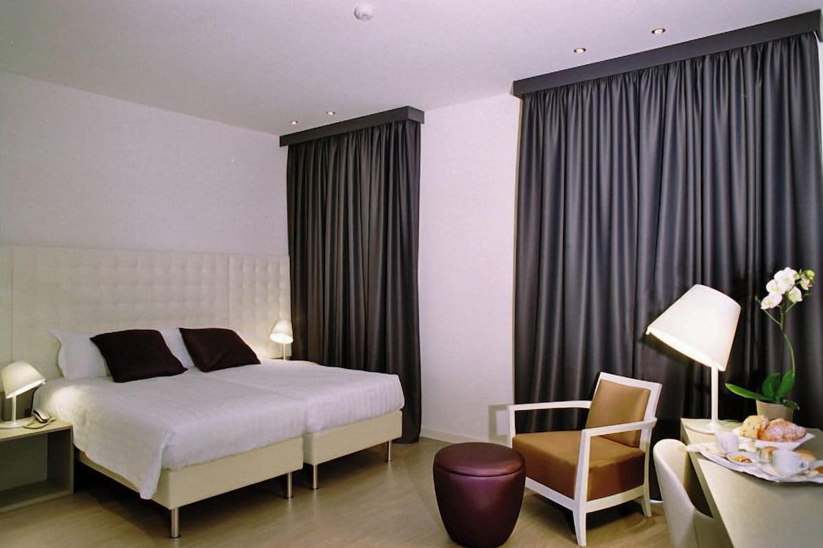 Una habitación de hotel moderna con una cama grande, un escritorio con silla, un taburete redondo pequeño y una orquídea en maceta.