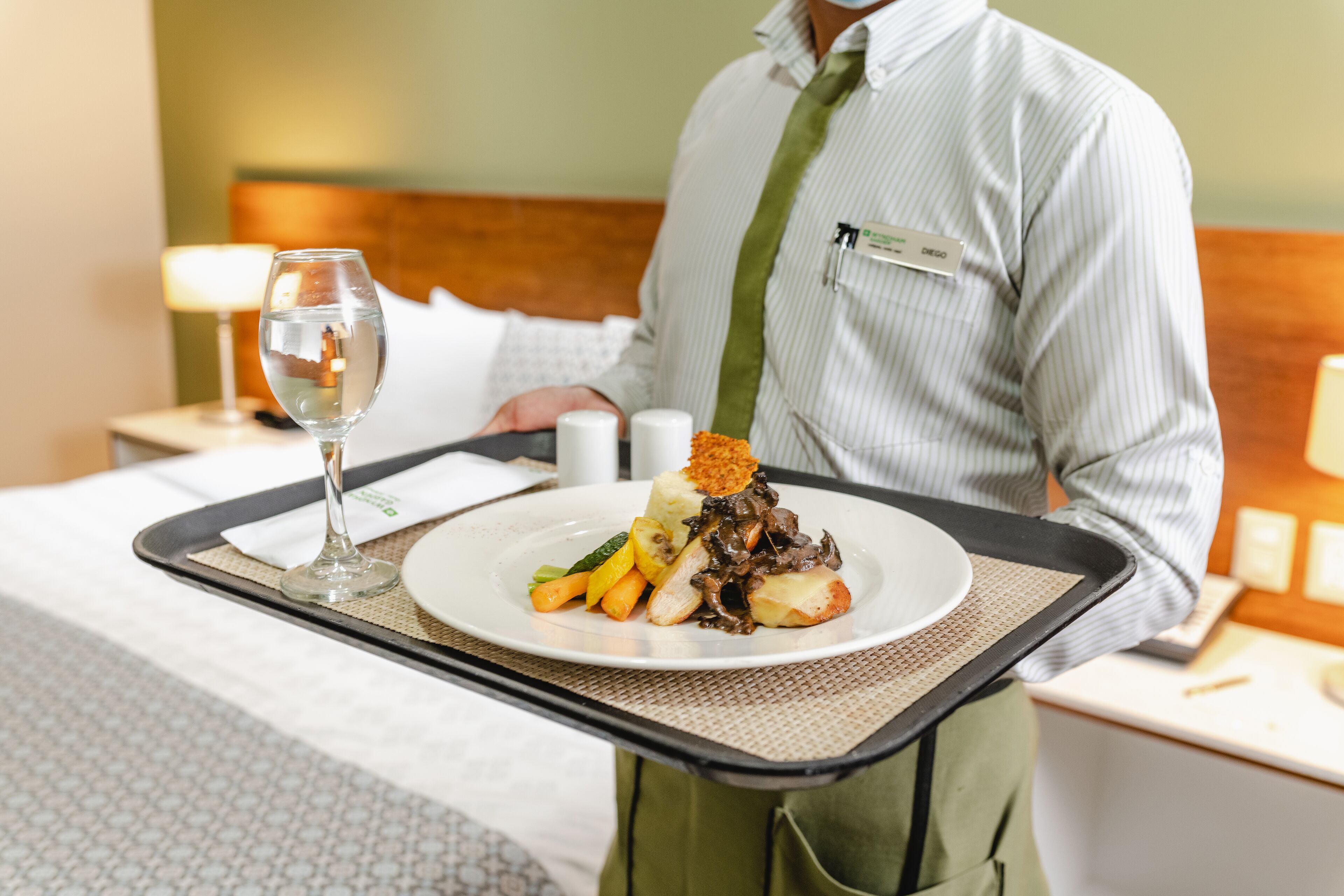 A hotel room with a neatly made bed, a bedside table with a lamp, and a tray with a plate of food and a glass of water.