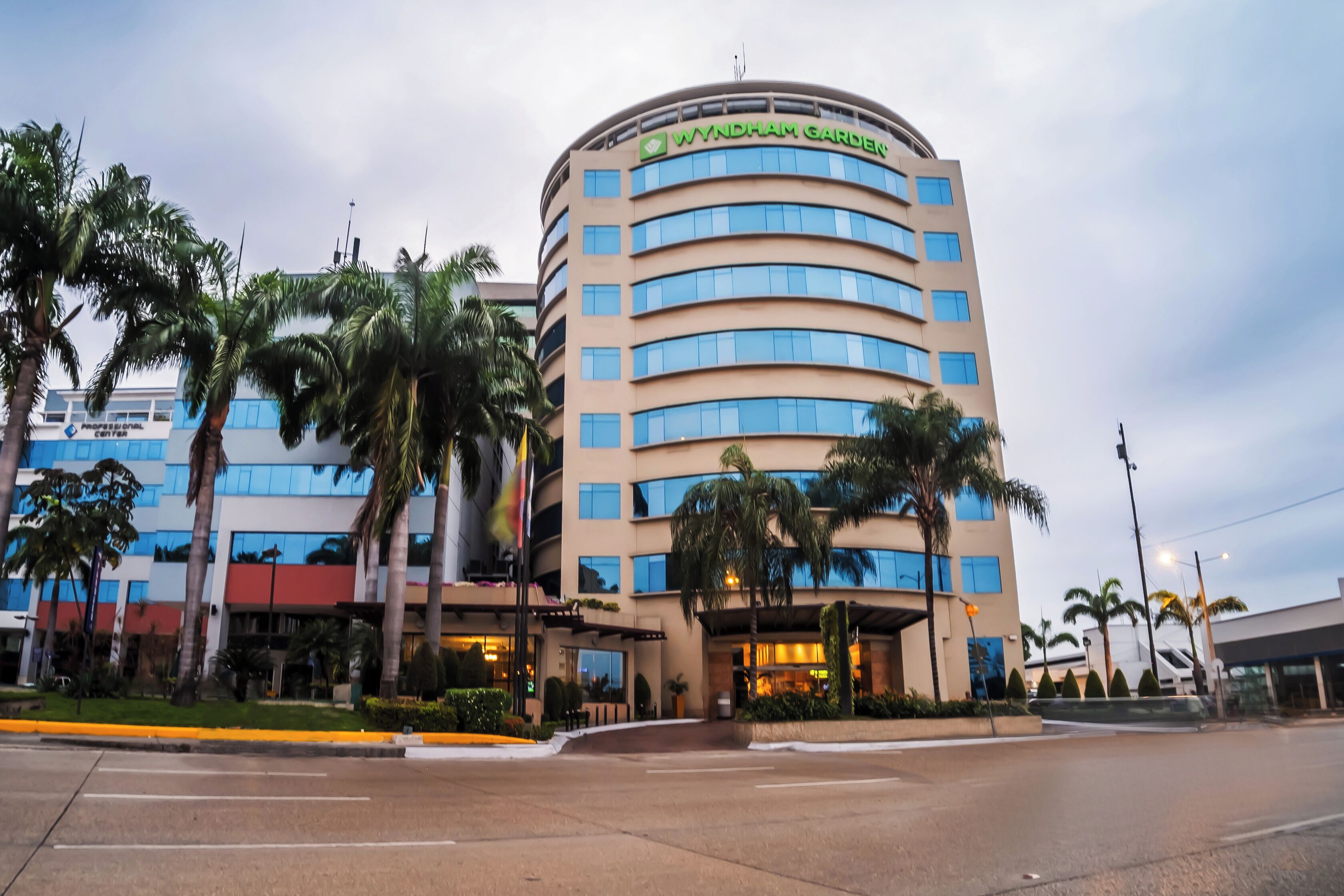 Un hotel de varios pisos con fachada curva, un llamativo letrero de 'Wyndham Garden' y una entrada ajardinada con palmeras.