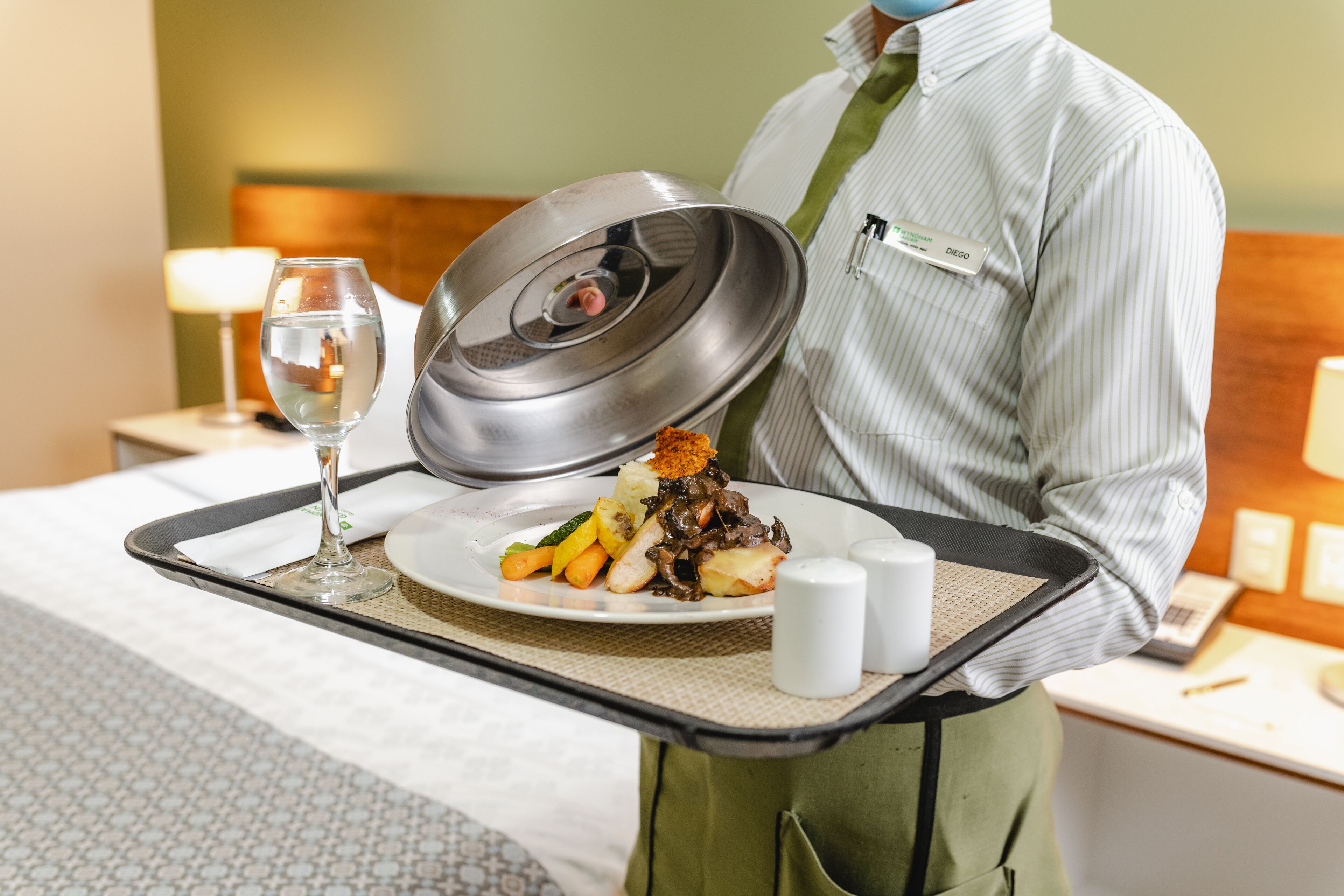 A hotel room with a neatly made bed, a bedside table with a lamp, and a tray with a plate of food and a glass of water.