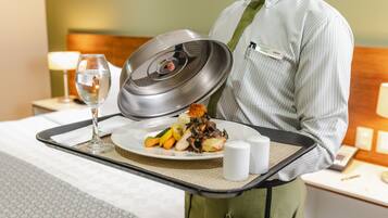 A hotel room with a neatly made bed, a bedside table with a lamp, and a tray with a plate of food and a glass of water.