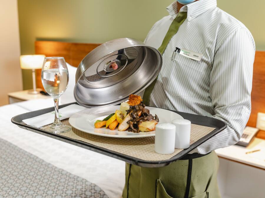 A hotel room with a neatly made bed, a bedside table with a lamp, and a tray with a plate of food and a glass of water.