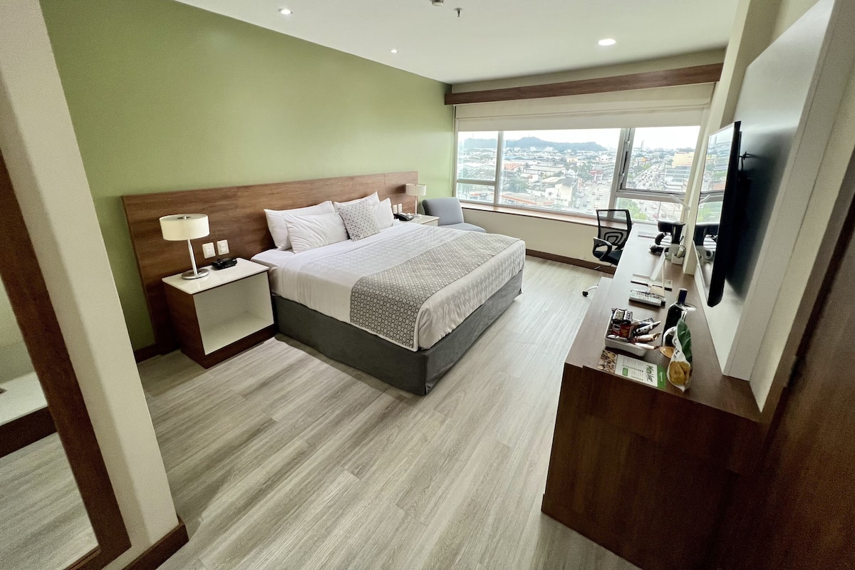 A hotel room with a large bed, a desk with a computer, a TV, and a view of the city through large windows.