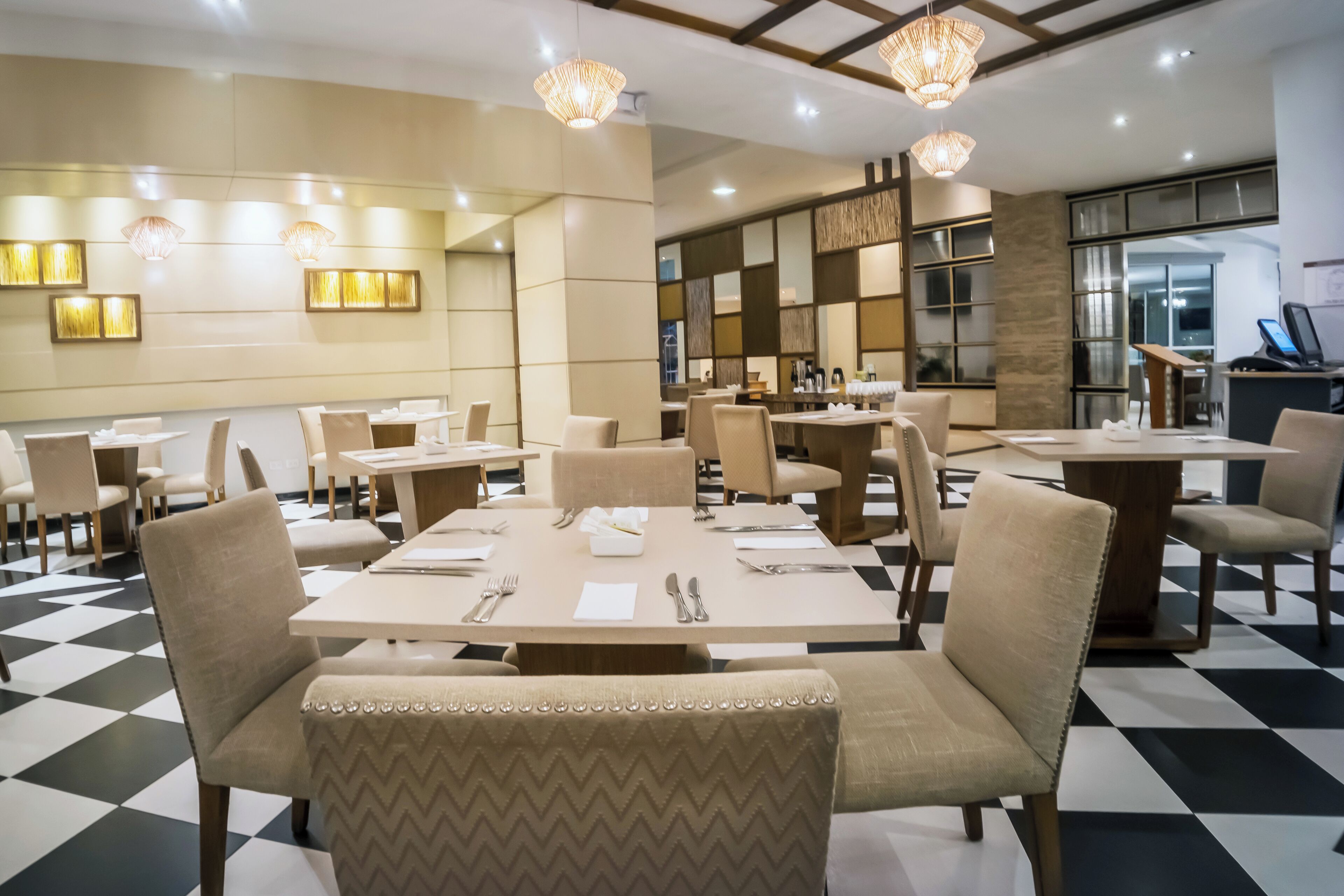A hotel dining area with multiple tables and chairs, a checkered floor, and modern lighting fixtures.