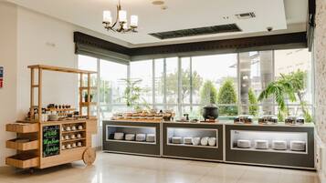 Una habitación de hotel con un ventanal grande, un carrito de madera con vajilla y una zona de kitchenette.
