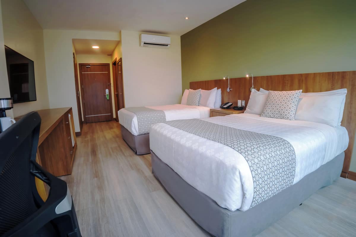 A hotel room with two beds, a desk, a chair, and an air conditioning unit.