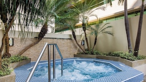 Un jacuzzi con escalera metálica, rodeado de palmeras y un muro de ladrillo.