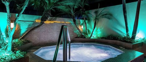 Un jacuzzi rodeado de palmeras y un muro de ladrillo, con una barandilla metálica en primer plano.