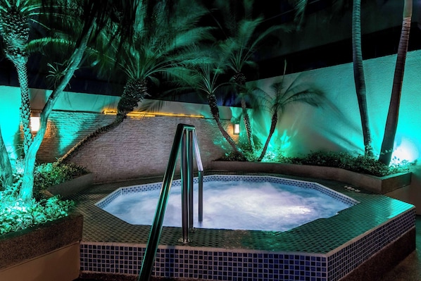 Un jacuzzi rodeado de palmeras y un muro de ladrillo, con una barandilla metálica en primer plano.