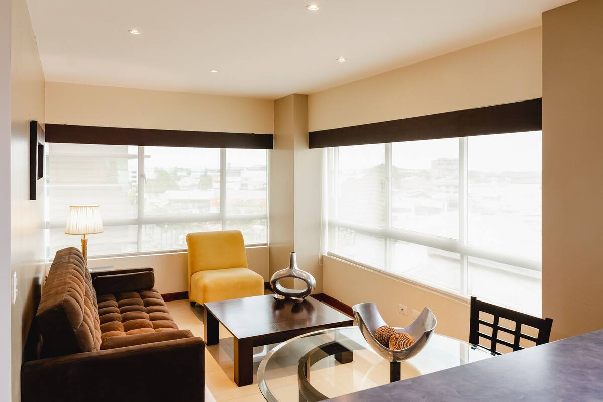 A modern living room with a sofa, a yellow chair, a coffee table, and a dining table with chairs. Large windows provide a view of the cityscape.