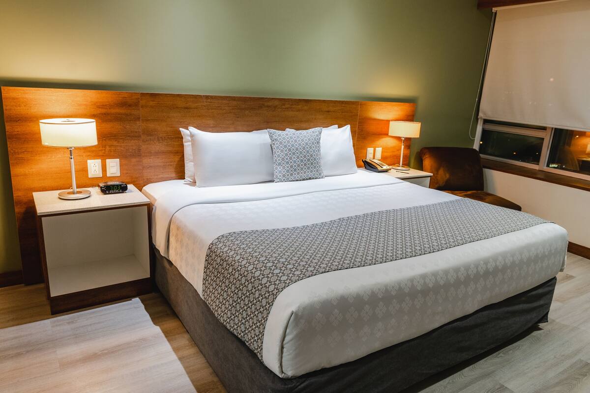 A neatly made bed with a patterned bedspread, two bedside lamps, a nightstand, and a window with a view.