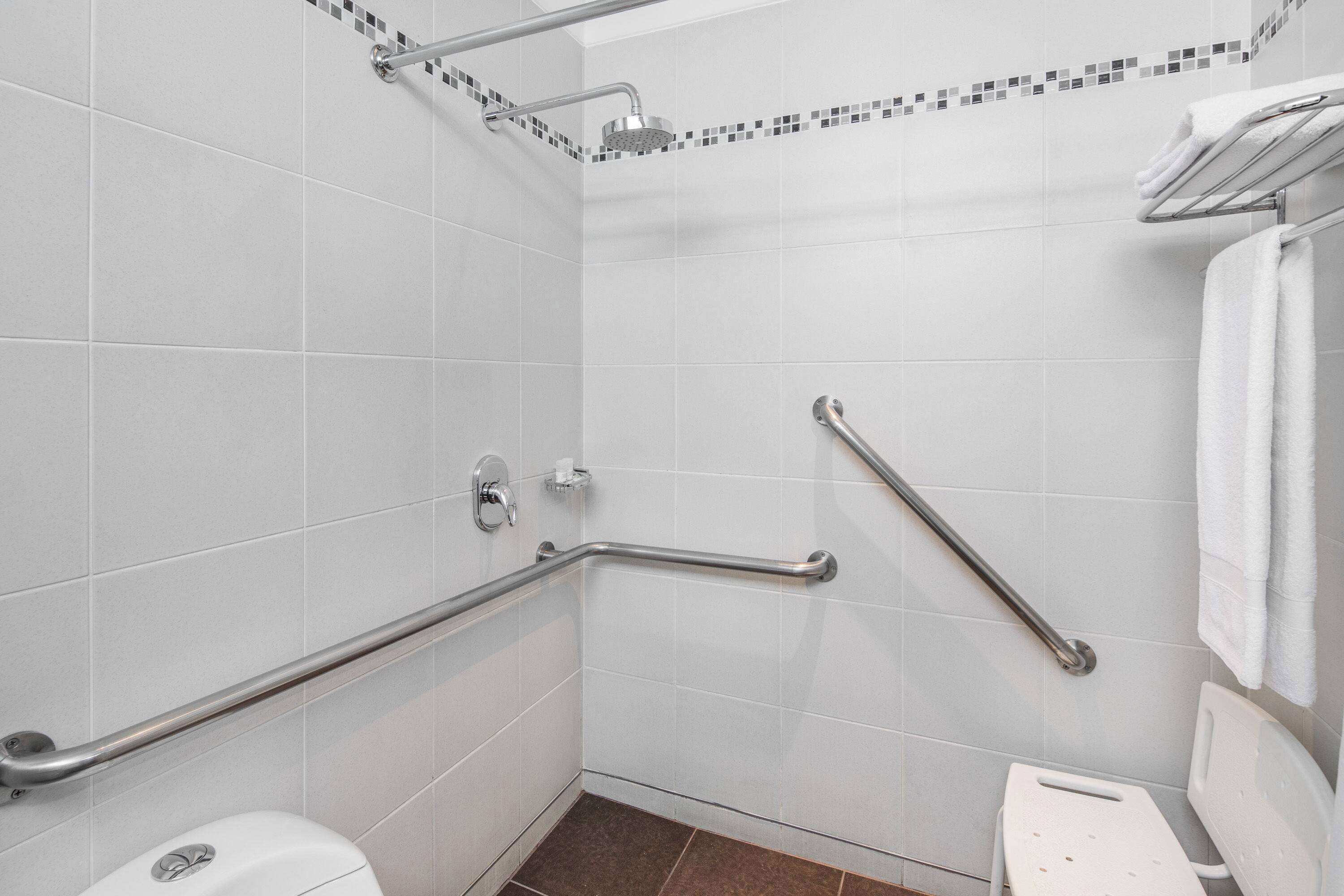 Un baño con azulejos blancos, una ducha con cabezal de ducha móvil, un barra de apoyo y un inodoro blanco.