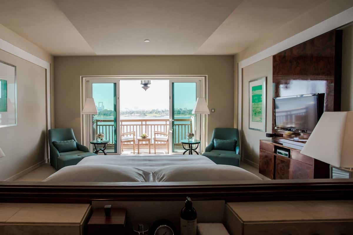 A hotel room with a large bed, two chairs, a TV, and a balcony with a view of the city.