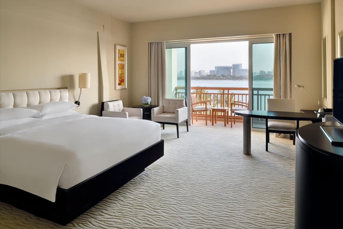 A hotel room with a large bed, a desk, a chair, a small table, and a view of a body of water.