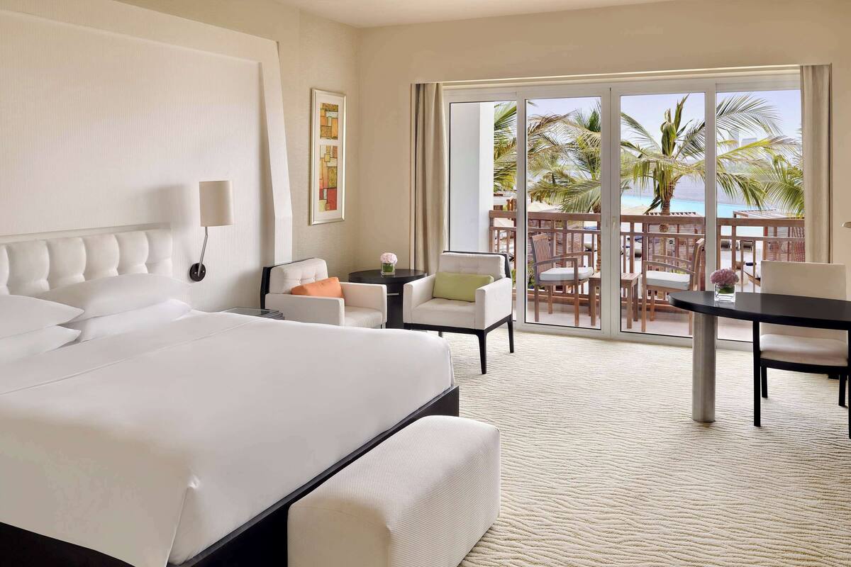 A hotel room with a large bed, a sofa, a small table, and a balcony with a view of the beach.
