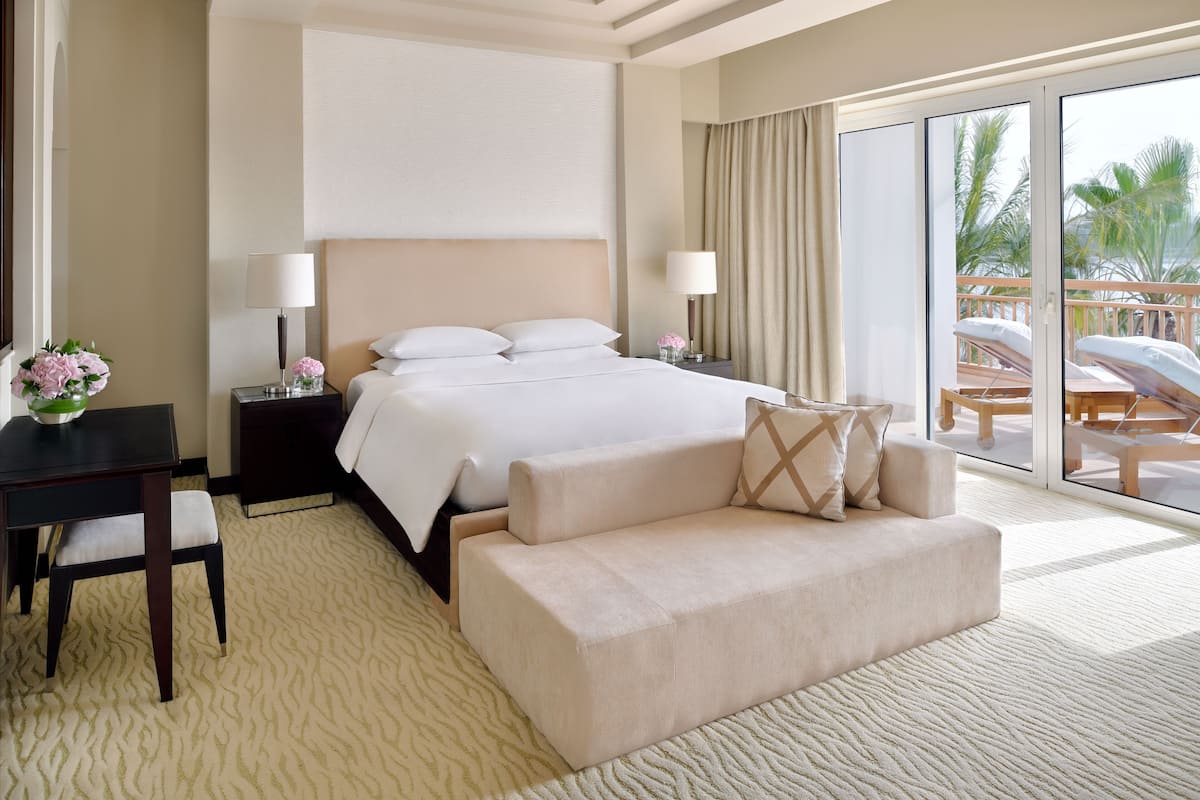 A hotel room with a large bed, a sofa, a desk, and a view of palm trees through the glass door.