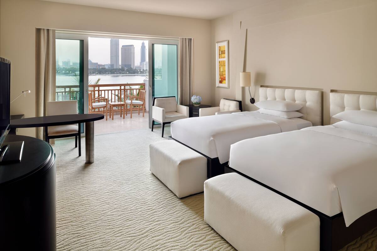 A hotel room with two beds, a desk, a chair, a TV, and a balcony with a view of the city.