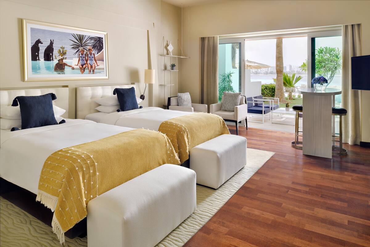 A hotel room with two beds, a sofa, a desk, and a large window offering a view of the city.