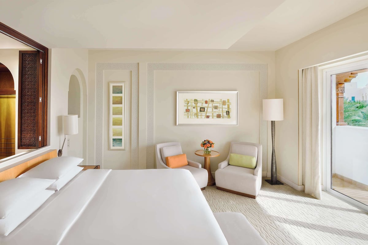 A hotel room with a large bed, two armchairs, a small table with flowers, a floor lamp, and a large window with a view.