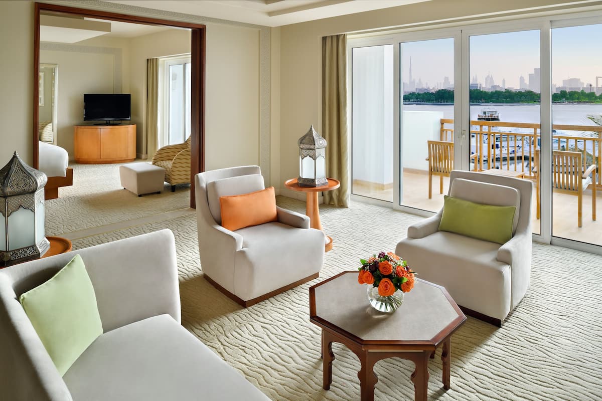 A hotel room with a large window, a flat-screen TV, a sofa, a chair, a small table with a vase of flowers, and a view of a city skyline.