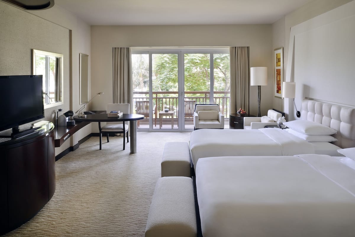 A hotel room with two beds, a desk, a TV, and a balcony with a view of trees.