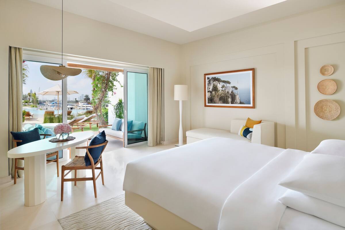 A modern hotel room with a large bed, a dining area with a table and chairs, a sofa, and a view of the pool and cityscape.