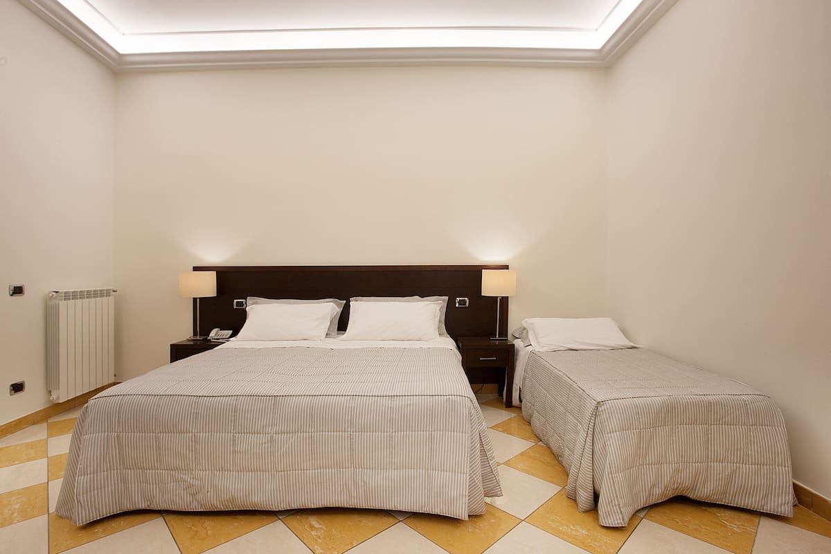 A hotel room with two beds, a skylight, a bedside table with a lamp, and a radiator.